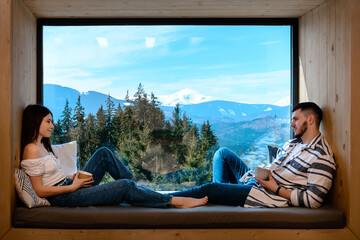 A young couple over a mug of coffee discussing everyday affairs, a guy and a girl sitting on the windowsill on a background of mountains and drinking coffee.