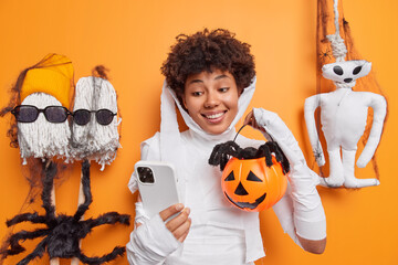 Happy smiling young woman wears ghost costume takes selfie of herself with carved pumpkin and...