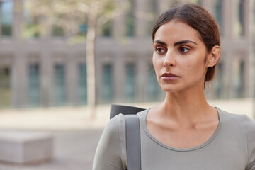 Horizontal shot of pensive good looking European sportswoman carries karemat poses against blurred background with empty space for your advertising content thinks how to keep fit and healthy