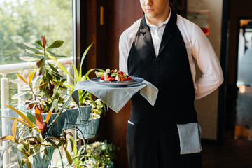 A young waiter in a stylish uniform stands with an exquisite dish on a tray near the table in a...