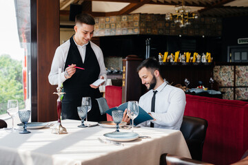 A young businessman in a fine restaurant examines the menu and makes an order to a young waiter in a stylish apron. Customer service.