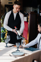 A young waiter in a stylish apron demonstrates and offers a fine wine to a client in a restaurant. Customer service.