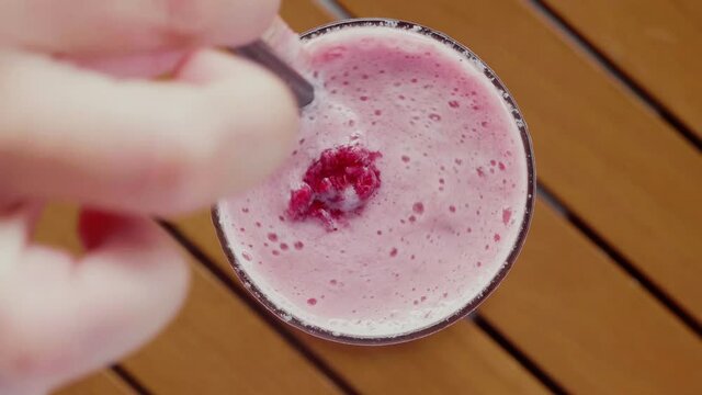Raspberry Smoothie in Glass. Close up of Woman Hand Stirs Fresh Pink Berry Vegetarian Drink or Strawberry Milkshake on Wooden Table. Healthy, Fitness Eating Concept. Top View