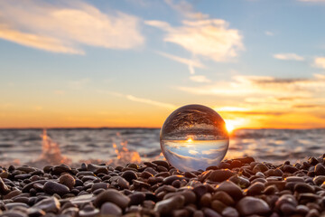 Obraz premium Clear glass ball sitting in colorful rocks at edge of Lake Michigan waves crash behind the scene at sunset. The sky is golden sun and blue sky