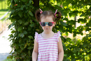 Little girl in a striped dress and sunglasses sadly looks at the camera against a blurred deciduous background