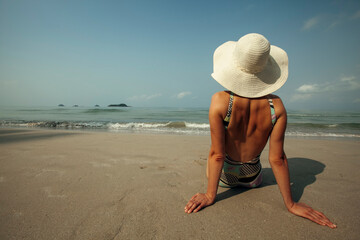 Fototapeta na wymiar A young woman is resting on the sea tropical beach. Sitting in a panama hat rear view.