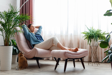 Happy Caucasian woman relax sit in pink chair in cozy home interior with red cat. Smiling young woman have a rest in armchair, enjoying comfort surrounded by home palm flowers Domestic cat pet.
