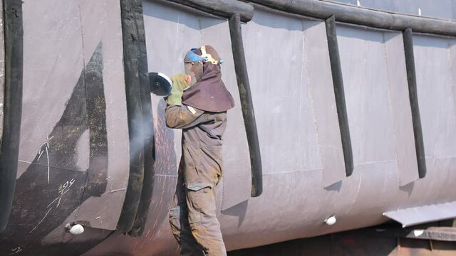 A worker with a safety helmet stands on a wooden plank working steel on a ship's hull with a angular grinder wheel in an industrial port in the city of Nijmegen in the Netherlands