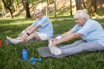 Positive senior man does head to knee exercise with wife sitting on meadow