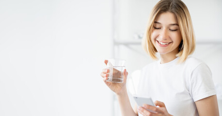Water refreshment. Healthy hydration. Morning nutrition. Cheerful woman with phone holding glass with liquid on defocused light copy space banner.