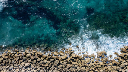 Aerial view from flying drone of crystal blue ocean water splashing on sea rocks.