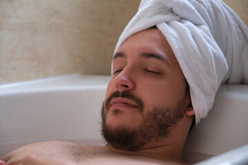 Young relaxed man with closed eyes lying in white bathtub filled with hot water.