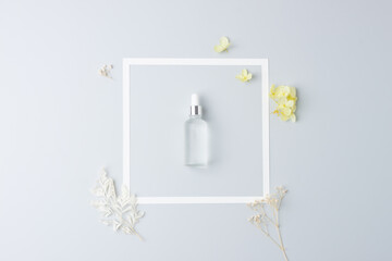 Cosmetic bottle with flowers and white frame on grey background. Flat lay, copy space