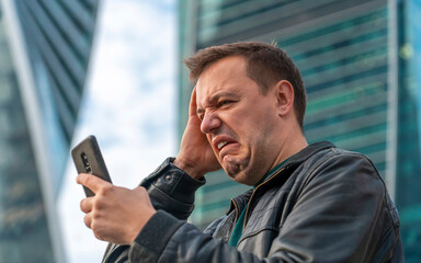 Sad millennial guy looks at phone screen in surprise and stands outside office building with city skyline in background. Disgusted and overwhelmed young man stares at screen of smartphone in cityscape