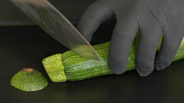 A cook slices a courgette lengthwise with a knife on a black board.  Close up Shot