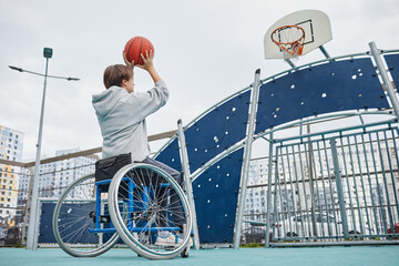 Rear view of woman with disability sitting in wheelchair throwing the ball up to the basket outdoors