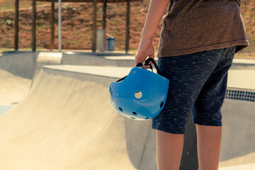 skater holding a blue helmet and a skateboard in front of bowl at skate park © Celso