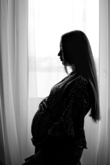 Pregnant woman silhouette photo. Pregnant female standing near the window. Side-view portrait of pregnant woman. Selective focus. Film grain.