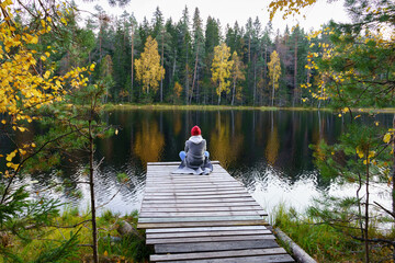Fototapeta na wymiar Woman sit on wooden dock relaxed enjoying landscape of autumn forest and lake. Back view of female relax on river pier in woods with colored yellow golden trees around. Nature beauty and inspiration