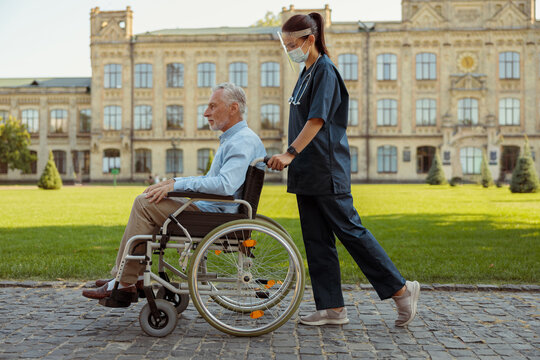 Full Length Shot Of Caring Young Nurse Wearing Face Shield And Mask Walking With Mature Man Patient In Wheelchair