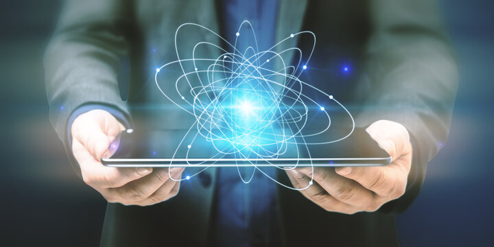 Close up of businessman hands holding tablet with abstract glowing atom hologram on dark background. Science and connection concept.