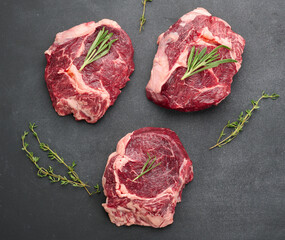 raw piece of beef ribeye with rosemary, thyme on a black table, top view