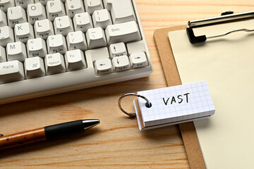 There is a word book with the word of VAST which is an abbreviation for Video Ad Serving Template on the desk with a pen and a keyboard.