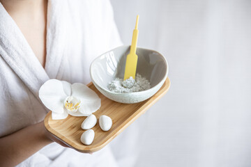 A young woman in a robe holds a bowl with a clay face mask.