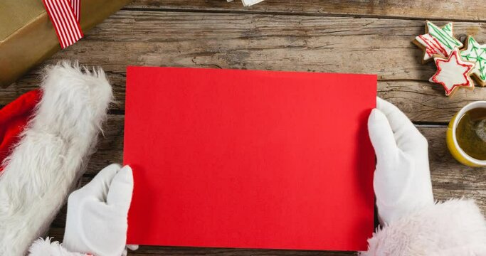 Animation of hands of santa claus holding red christmas card with copy space over wooden background