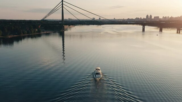 Modern white motorboat yacht cruise sunset river. Cityscape buildings in background. Orange sun over a sailboat and calm water. Tourist tour excursion, voyage, cruise. Aerial drone view. Kyiv, Ukraine