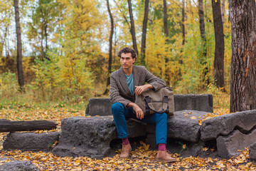 Tall handsome man with a bag sitting on the rocks in the autumn forest