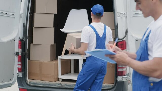 Two young workers of removal company are loading boxes and furniture into a minibus