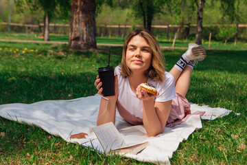 Young romantic woman laying on the grass in park with open book, cake and cup of coffee to go