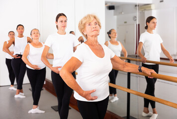 Group of ballerina standing with one hand on barre and reaching over hand in bright fitness room