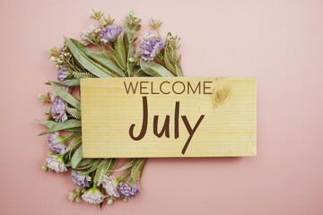 Welcome July text on wooden board with flowers frame on pink background