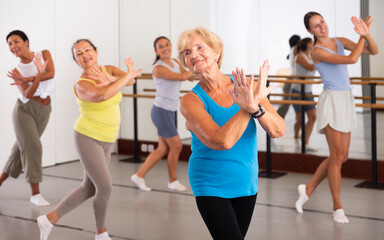 Fototapeta na wymiar Portrait of smiling mature woman practicing ballet dance moves during group class in choreographic studio