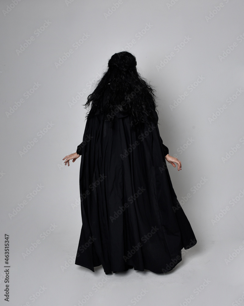 Canvas Prints Full length portrait of dark haired woman wearing  black victorian witch costume with  cloak and pointy hat.  standing pose, back view,  with  gestural hand movements,  against studio background.