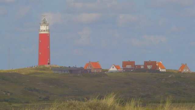 Steady Tele shot of Waving Marram grass in the foreground. In the distance we see a lighthouse in the dunes together with some builings. The sky is blue with seagulls and beautiful clouds passing by.