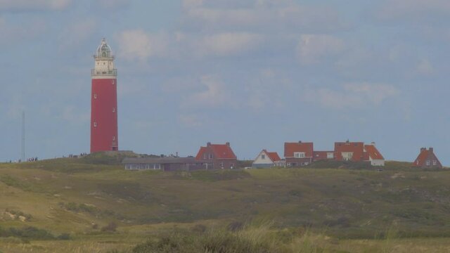 Steady Tele shot of Marram grass in the foreground. In the distance a lighthouse in the dunes together with some builings. first in the shadow, later in the sun. The sky is blue with clouds passing by