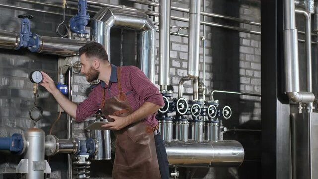 Young male brewer wearing a leather apron supervise the process of beer fermentation at a modern brewery factory