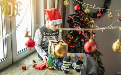 Little boy in a red Santa hat is sitting at the window next to a Christmas tree in a room decorated for the holiday and holding a gift box in his hands. Concept of New Year atmosphere and festive mood