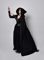 Naklejka premium Full length portrait of dark haired woman wearing black victorian witch costume with a flowing cloak. standing pose, with gestural hand movements, against studio background.