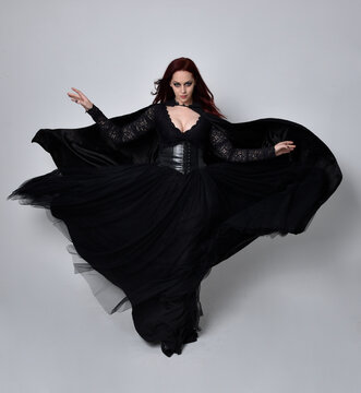 Full length portrait of dark haired woman wearing  black victorian witch costume with a flowing  cloak.  standing pose, with  gestural hand movements,  against studio background.