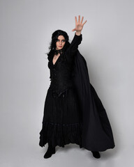 Full length portrait of dark haired woman wearing  black victorian witch costume with a flowing  cloak.  standing pose, with  gestural hand movements,  against studio background.