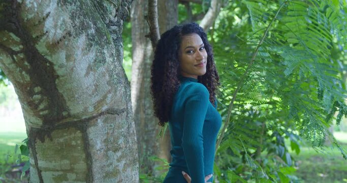 An east indian model with curly hair stands next to a large tree trunk in green dress