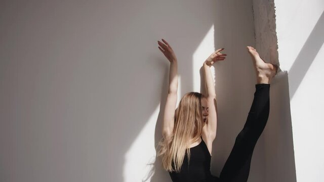 A beautiful young girl stands on a stretch in a longitudinal twine against the wall and straightens her long hair