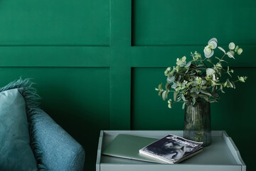 Stylish glass vase with eucalyptus on table near green wall