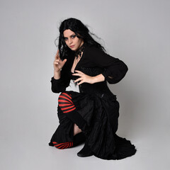 Full length portrait of dark haired woman wearing  black victorian witch costume  sitting pose, with  gestural hand movements,  against studio background.