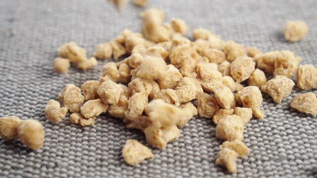 Textured chunks of organic dehydrated soybeans fall on a rough jute surface in slow motion close up. Vegetable soy breakfast flakes