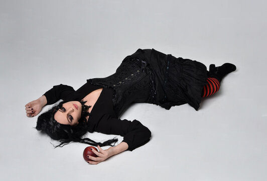Full length portrait of dark haired woman wearing  black victorian witch costume,  lying on the floor as is dead for asleep, isolated on studio background.